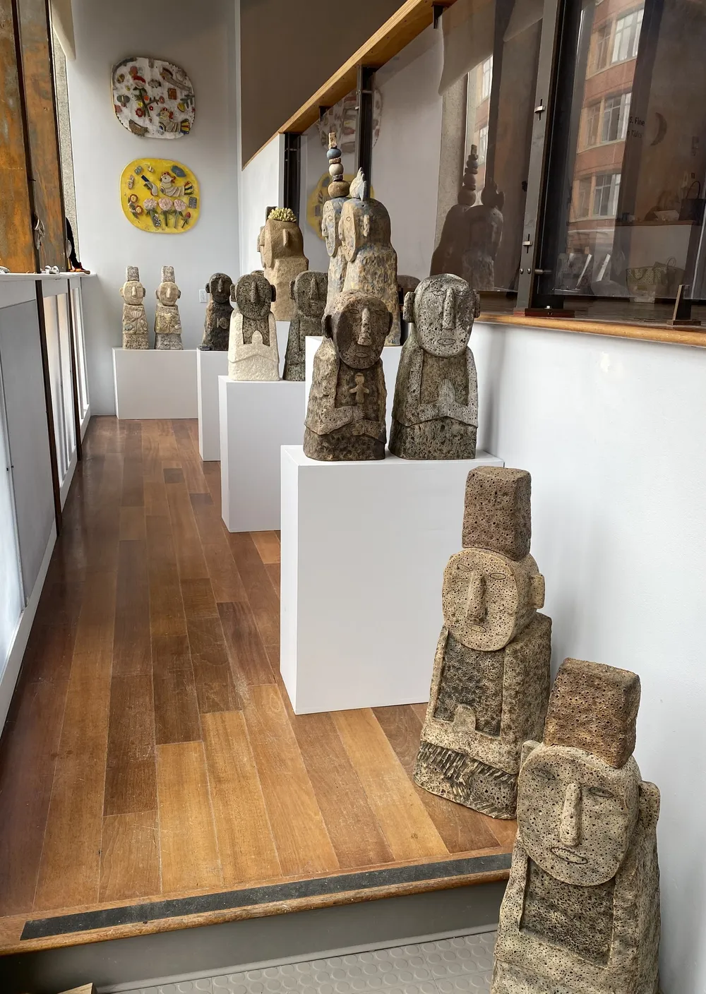 Family of All Things in the Launch Pad corridor at Boston Sculptors Gallery: rows of clay guardian figures on white plinths receding down a wood floor, with two round painted wall panels, one yellow, at the far end.