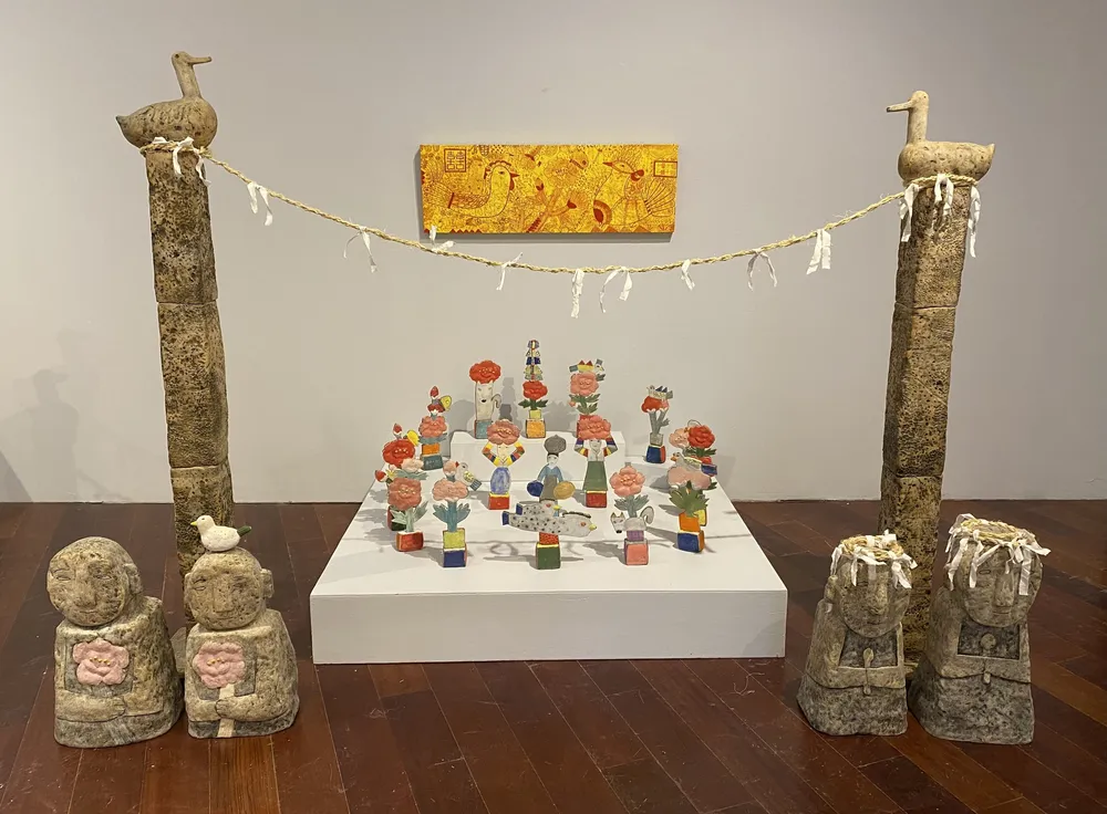 Peonies for My Mother seen head-on: a white platform crowded with small clay figures carrying peonies, flanked by two clay sotdae poles that carry a straw rope hung with white cloth strips, four guardian figures on the floor and a long yellow minhwa panel on the wall above.