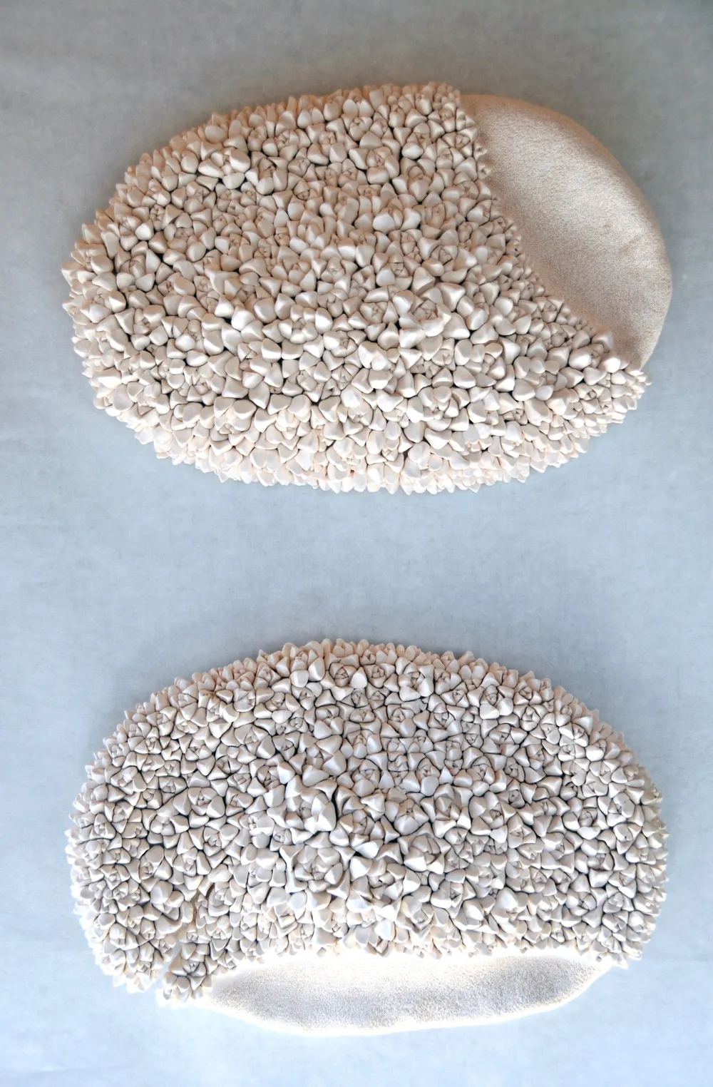 Two oval echeveria wall pieces photographed one above the other — each a mass of small white porcelain nubs, the upper finished with a smooth sanded lobe at the right, the lower with a sanded band along the bottom.