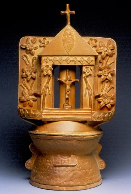 An ochre censer shaped like a shrine facade — a cross at the peak, a gabled roof, a crucifix in the central arch and flowering vines on the wings — set on a domed footed bowl.