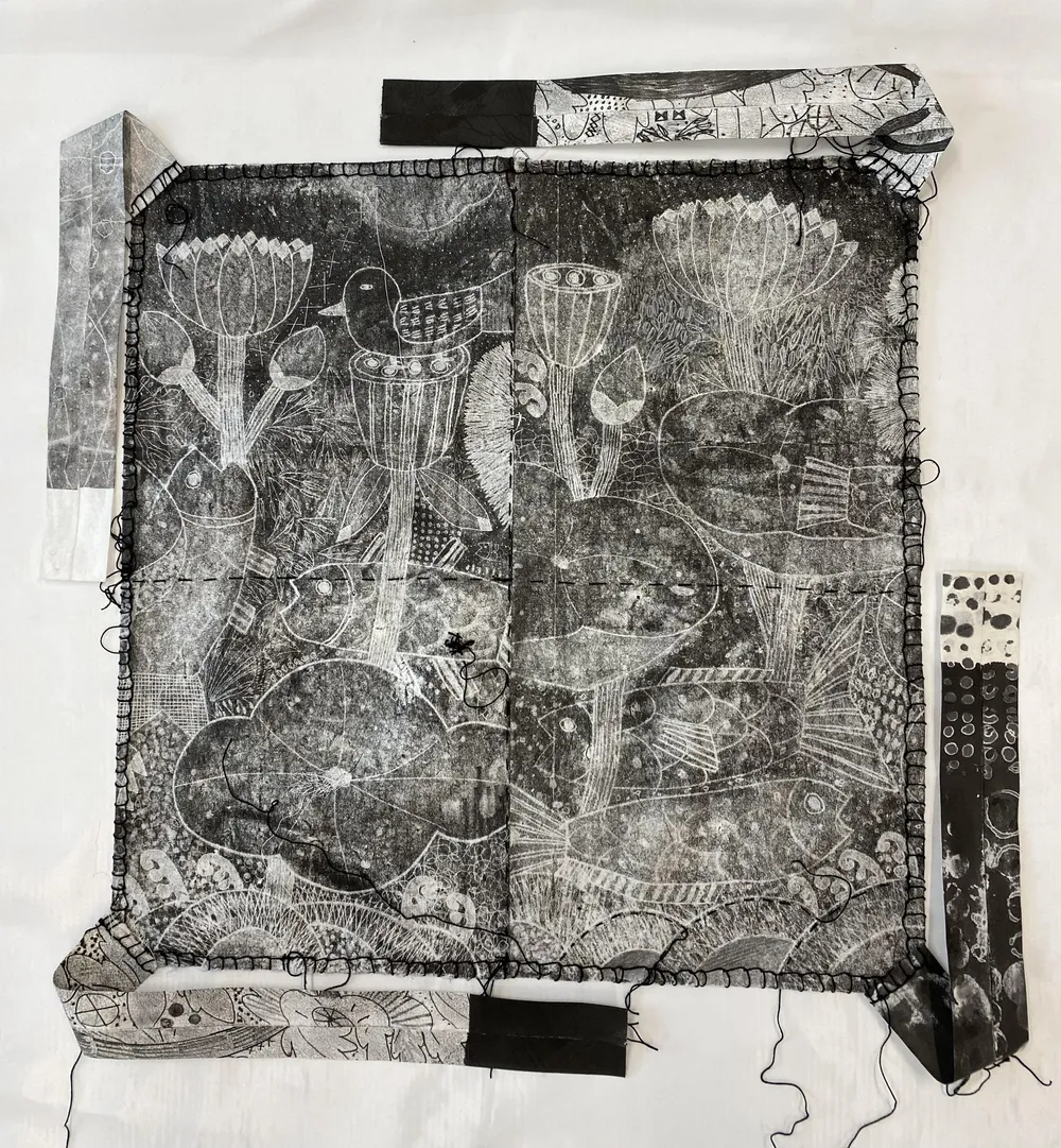 A black-and-white monoprint bojagi on cloth — lotus pods, a perched bird and a fish — machine-stitched with a black border, loose thread ends and four printed strips running off the edges, laid flat on white paper.