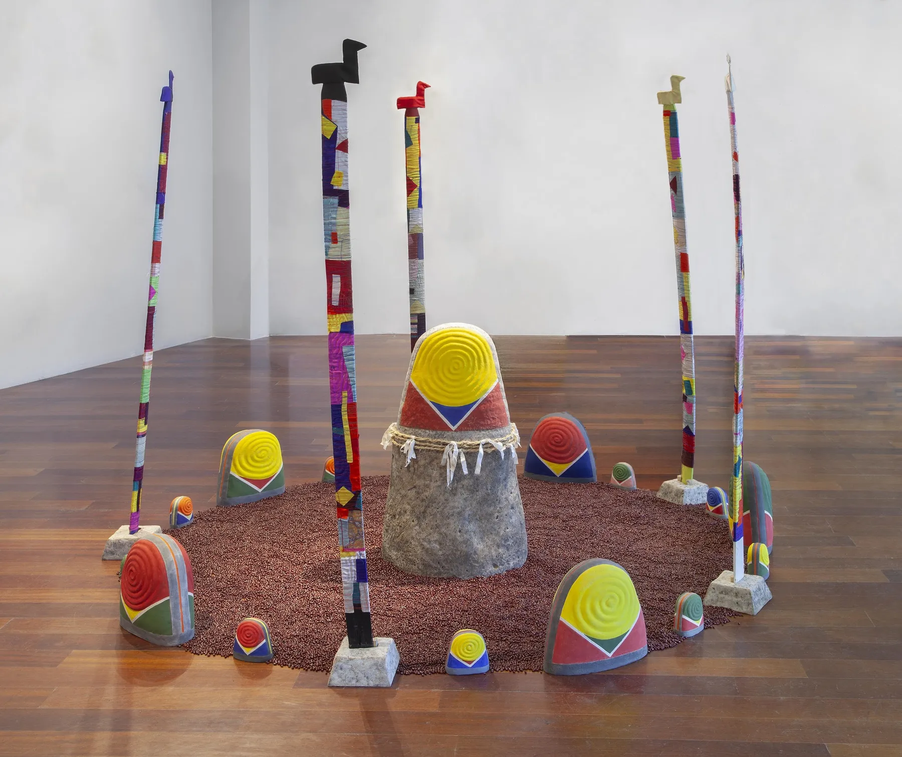 Sanctuary, 2025: a large stone-textured thimble form at the centre of a circle of small painted thimble domes, ringed with red adzuki beans and surrounded by silk-wrapped picket-post sotdae topped with ceramic birds.