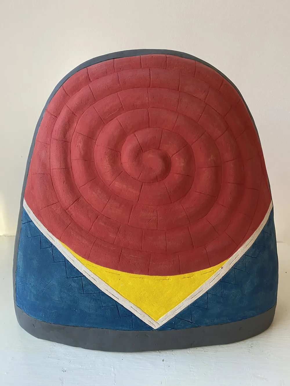Thimble 4, a large clay thimble dome: a red concentric spiral cap over a blue body with a yellow chevron and a dark grey foot band, leaning against a white wall.