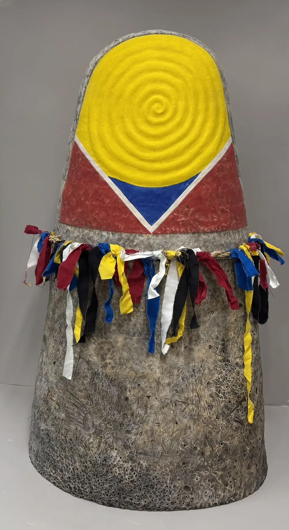 Ceramic Thimble, a single large thimble form against a grey wall: a yellow concentric spiral over red with a blue triangle, its stone-textured lower body ringed with tied strips of obangsaek cloth in red, yellow, blue, white and black.