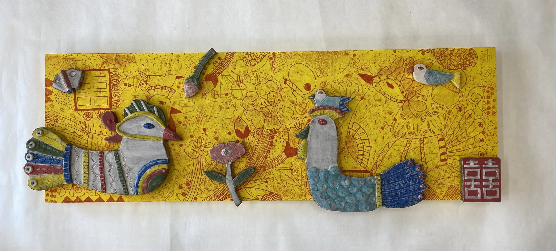 A long horizontal talisman panel on a yellow ink-drawn ground, hung with ceramic pieces — two crested birds, small fish, a flower and a red double-happiness seal at the right.